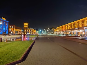 A nighttime scene of a city square with illuminated buildings featuring bright blue and warm orange lighting. The area is lined with well-kept green grass and colorful fountains creating a lively atmosphere. The cobblestone walkway suggests a modern yet historic architectural style.
