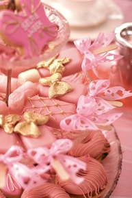 An assortment of pink-themed desserts arranged on a tiered glass stand. The treats include heart-shaped pink cookies adorned with golden butterflies and bows, complemented by delicate pink ribbons. Some desserts are set on sticks, resembling cake pops, and feature intricate pink icing designs. A hint of tea or coffee service is visible in the background.