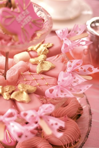 An assortment of pink-themed desserts arranged on a tiered glass stand. The treats include heart-shaped pink cookies adorned with golden butterflies and bows, complemented by delicate pink ribbons. Some desserts are set on sticks, resembling cake pops, and feature intricate pink icing designs. A hint of tea or coffee service is visible in the background.
