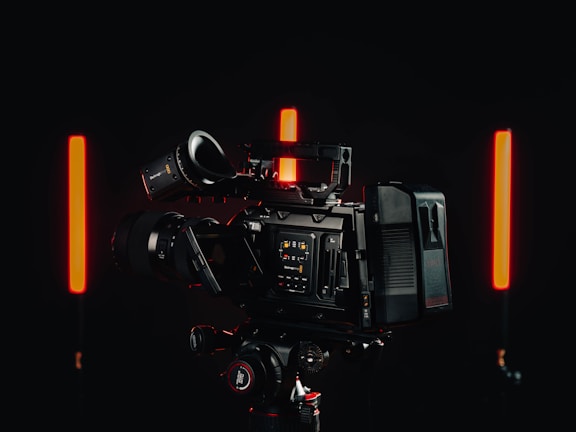 A professional video camera with a detailed control interface is positioned in a dimly lit environment. Two vertical red lights are on either side of the camera, casting a soft glow against the dark background.