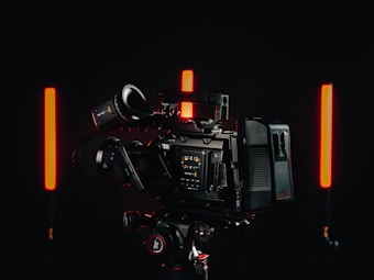 A professional video camera with a detailed control interface is positioned in a dimly lit environment. Two vertical red lights are on either side of the camera, casting a soft glow against the dark background.