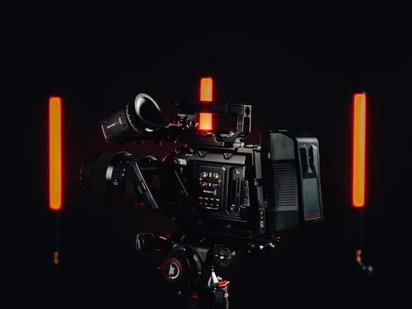 A professional video camera with a detailed control interface is positioned in a dimly lit environment. Two vertical red lights are on either side of the camera, casting a soft glow against the dark background.