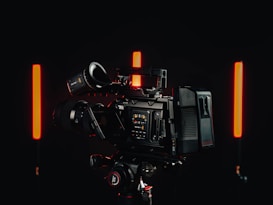 A professional video camera with a detailed control interface is positioned in a dimly lit environment. Two vertical red lights are on either side of the camera, casting a soft glow against the dark background.