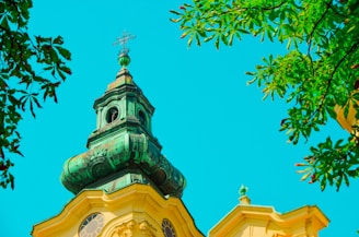 A vibrant church steeple with a patina green tower roof set against a bright, clear sky. Surrounding the steeple, lush green leaves from nearby trees frame the scene.