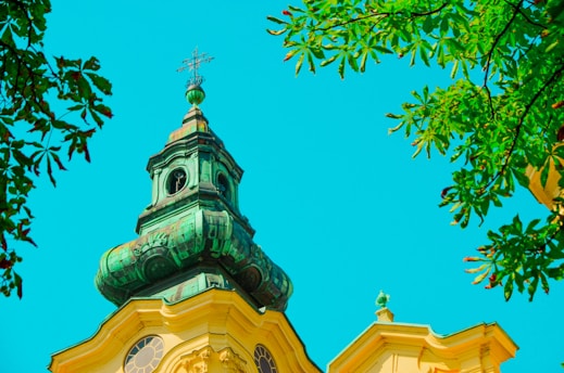 A vibrant church steeple with a patina green tower roof set against a bright, clear sky. Surrounding the steeple, lush green leaves from nearby trees frame the scene.