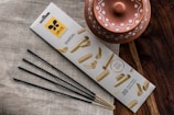 A package of sandal-scented incense sticks is placed on a textured fabric surface. The packaging features an attractive design with images of sandalwood. Next to it, a rustic terracotta pot with decorative white patterns is visible. The scene is set against a wooden surface, adding an earthy aesthetic.