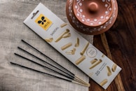 A package of sandal-scented incense sticks is placed on a textured fabric surface. The packaging features an attractive design with images of sandalwood. Next to it, a rustic terracotta pot with decorative white patterns is visible. The scene is set against a wooden surface, adding an earthy aesthetic.