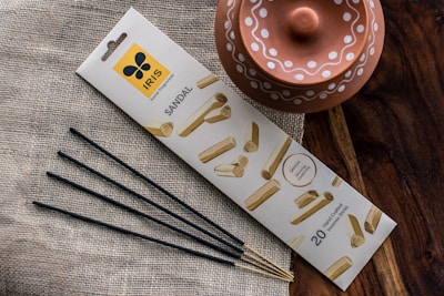 A package of sandal-scented incense sticks is placed on a textured fabric surface. The packaging features an attractive design with images of sandalwood. Next to it, a rustic terracotta pot with decorative white patterns is visible. The scene is set against a wooden surface, adding an earthy aesthetic.