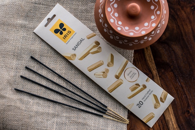 A package of sandal-scented incense sticks is placed on a textured fabric surface. The packaging features an attractive design with images of sandalwood. Next to it, a rustic terracotta pot with decorative white patterns is visible. The scene is set against a wooden surface, adding an earthy aesthetic.