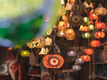 Colorful mosaic lanterns hanging in a market setting, displaying intricate patterns and vibrant hues. The scene is rich with decorative glasswork, featuring various designs and a warm, inviting glow.