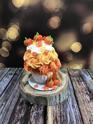 A decorative cupcake is placed on a round wooden slab. The cupcake is adorned with orange and white swirls of frosting, topped with small fondant pumpkins and autumn leaves. The background features a warm, blurred bokeh effect with soft golden lights.