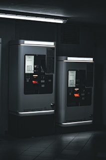 Two parking ticket machines are situated in a dimly-lit area, with a fluorescent light fixture above casting a cool, stark glow. The machines display various informational panels and illuminated screens. The ambient setting appears industrial, with tiled flooring and a dark background.