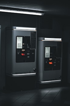Two parking ticket machines are situated in a dimly-lit area, with a fluorescent light fixture above casting a cool, stark glow. The machines display various informational panels and illuminated screens. The ambient setting appears industrial, with tiled flooring and a dark background.