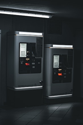 Two parking ticket machines are situated in a dimly-lit area, with a fluorescent light fixture above casting a cool, stark glow. The machines display various informational panels and illuminated screens. The ambient setting appears industrial, with tiled flooring and a dark background.