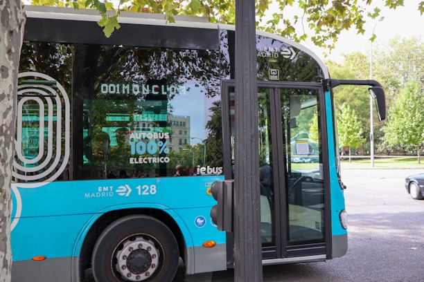 A modern electric bus with a blue and silver color scheme is parked on a city street. The side of the bus has markings indicating it is 100% electric, along with the logo of EMT Madrid. The scene is surrounded by trees and there is a building in the background. Part of a tree and a streetlamp are visible in the foreground.