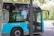 A modern electric bus with a blue and silver color scheme is parked on a city street. The side of the bus has markings indicating it is 100% electric, along with the logo of EMT Madrid. The scene is surrounded by trees and there is a building in the background. Part of a tree and a streetlamp are visible in the foreground.