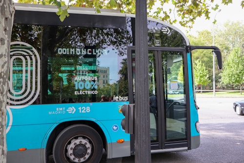 A modern electric bus with a blue and silver color scheme is parked on a city street. The side of the bus has markings indicating it is 100% electric, along with the logo of EMT Madrid. The scene is surrounded by trees and there is a building in the background. Part of a tree and a streetlamp are visible in the foreground.