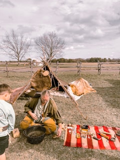 A person wearing rustic, historical clothing is seated on the grass, engaging in a traditional activity with various tools and items spread out on a red and white striped blanket. Nearby, a simple structure resembling a small tipi made with animal hides is set up. Another individual, possibly a child, stands watching. The setting is outdoors, with fenced open fields and bare trees in the background under a cloudy sky.
