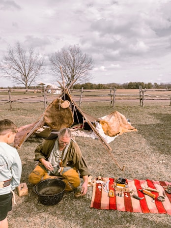 A person wearing rustic, historical clothing is seated on the grass, engaging in a traditional activity with various tools and items spread out on a red and white striped blanket. Nearby, a simple structure resembling a small tipi made with animal hides is set up. Another individual, possibly a child, stands watching. The setting is outdoors, with fenced open fields and bare trees in the background under a cloudy sky.