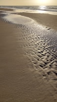 Golden sand beach with intricate patterns formed by the receding tide as the sun reflects off the wet sand and distant ocean.