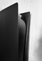 A close-up view of a sleek, modern gaming console standing vertically against a light wooden wall. The console has a two-tone color scheme, primarily black and white, with the brand name 'SONY' visible on the top right side.