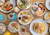 A table filled with a variety of brunch dishes, including pancakes with fruit and syrup, a burger with fries, a salad with poached eggs, a cappuccino, and a teapot. The presentation is artistic, with garnishes and sauces carefully arranged.