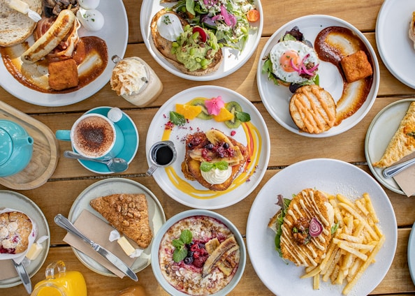 A table filled with a variety of brunch dishes, including pancakes with fruit and syrup, a burger with fries, a salad with poached eggs, a cappuccino, and a teapot. The presentation is artistic, with garnishes and sauces carefully arranged.