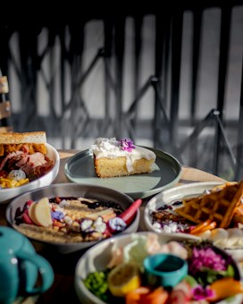 A selection of appetizing dishes is artfully arranged on a wooden table. The centerpiece is a slice of cake topped with white icing and a purple edible flower. Surrounding it are various dishes including a plate with waffles, a bowl of assorted fruits and vegetables, and a bowl containing meat and what appears to be macaroni and cheese. The setting is well-lit by natural light, casting soft shadows and highlighting the vibrant colors of the food.