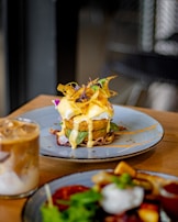 A gourmet dish is plated elegantly on a round ceramic plate. The dish consists of a golden-brown fried patty topped with white sauce, garnished with vibrant green leaves and crispy orange strips. A creamy, iced coffee drink is placed beside it on the wooden table.