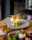 A gourmet dish is plated elegantly on a round ceramic plate. The dish consists of a golden-brown fried patty topped with white sauce, garnished with vibrant green leaves and crispy orange strips. A creamy, iced coffee drink is placed beside it on the wooden table.