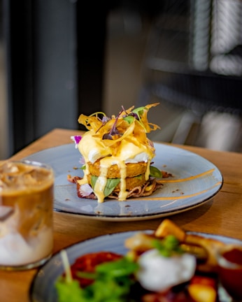 A gourmet dish is plated elegantly on a round ceramic plate. The dish consists of a golden-brown fried patty topped with white sauce, garnished with vibrant green leaves and crispy orange strips. A creamy, iced coffee drink is placed beside it on the wooden table.