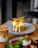 A gourmet dish is plated elegantly on a round ceramic plate. The dish consists of a golden-brown fried patty topped with white sauce, garnished with vibrant green leaves and crispy orange strips. A creamy, iced coffee drink is placed beside it on the wooden table.