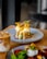 A gourmet dish is plated elegantly on a round ceramic plate. The dish consists of a golden-brown fried patty topped with white sauce, garnished with vibrant green leaves and crispy orange strips. A creamy, iced coffee drink is placed beside it on the wooden table.