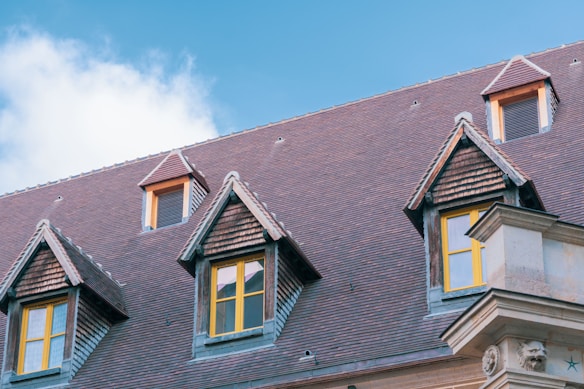 A building with a steeply pitched roof covered in red tiles features multiple gabled dormer windows with yellow frames. The sky is clear and blue with a few clouds visible.