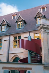 A traditional building with steeply pitched roofs featuring dormer windows, stone walls, and yellow-framed windows. A modern red spiral staircase curves in front of the building, adding a contemporary element to the classical architecture.