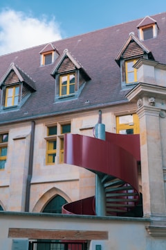 A traditional building with steeply pitched roofs featuring dormer windows, stone walls, and yellow-framed windows. A modern red spiral staircase curves in front of the building, adding a contemporary element to the classical architecture.