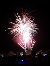 A vibrant display of fireworks illuminates the night sky, with bright streaks of light and colorful bursts. Below, a crowd of silhouettes watches the spectacle, surrounded by trees and some visible tents or structures. The atmosphere is festive and lively, capturing a moment of celebration.