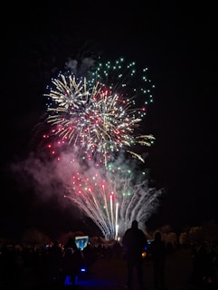 Bright multicolored fireworks burst against the night sky, with streaks of red, green, and blue illuminating the darkness. In the foreground, silhouettes of people gather to watch the spectacle, creating a lively and festive atmosphere.