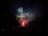 Vibrant fireworks light up the night sky with bursts of green, red, and blue hues. Silhouetted figures on the ground watch the display, adding a sense of scale and wonder to the scene. Plumes of smoke drift across the dark backdrop, enhancing the dramatic effect of the fireworks.