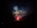 Vibrant fireworks light up the night sky with bursts of green, red, and blue hues. Silhouetted figures on the ground watch the display, adding a sense of scale and wonder to the scene. Plumes of smoke drift across the dark backdrop, enhancing the dramatic effect of the fireworks.