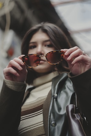 A stylish woman trying on oversized tortoiseshell sunglasses beside a bright window.