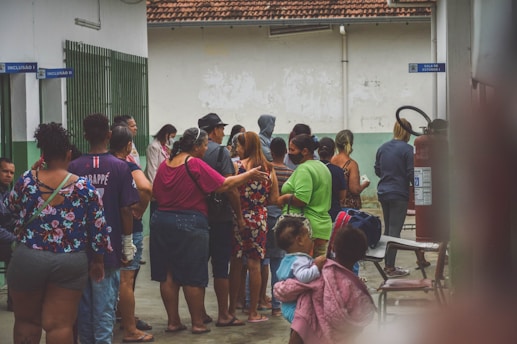 A group of people is gathered in an outdoor area, possibly near a building with signs and a fire extinguisher visible. The individuals are of various ages and ethnicities, dressed in casual clothing, some carrying bags. The background includes a green and white wall with sections peeling and a tiled roof above.