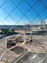 An airplane from Viva Aerobus, predominantly white with green accents, is parked at an airport gate. Ground crew members wearing orange vests are standing near luggage carts and equipment. The scene is viewed through a chain-link fence, and the sky is clear and blue.