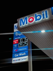 A gas station sign is illuminated at night, featuring the Mobil logo prominently. Various services are advertised, including a car wash and Mobil Mart. The prices of different types of fuel are displayed digitally, with labels for Super Plus and another grade. The backdrop of the image is predominantly dark, highlighting the brightly lit sign.