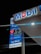 A gas station sign is illuminated at night, featuring the Mobil logo prominently. Various services are advertised, including a car wash and Mobil Mart. The prices of different types of fuel are displayed digitally, with labels for Super Plus and another grade. The backdrop of the image is predominantly dark, highlighting the brightly lit sign.