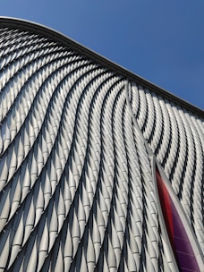 A modern architectural facade featuring a series of repeating, undulating metal panels that create a textured and dynamic surface. The surface reflects light and casts soft shadows, enhancing its three-dimensional appearance. In the background, a clear blue sky provides a stark contrast to the intricate patterns of the metal.