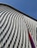 A modern architectural facade featuring a series of repeating, undulating metal panels that create a textured and dynamic surface. The surface reflects light and casts soft shadows, enhancing its three-dimensional appearance. In the background, a clear blue sky provides a stark contrast to the intricate patterns of the metal.