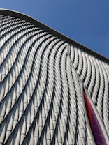 A modern architectural facade featuring a series of repeating, undulating metal panels that create a textured and dynamic surface. The surface reflects light and casts soft shadows, enhancing its three-dimensional appearance. In the background, a clear blue sky provides a stark contrast to the intricate patterns of the metal.