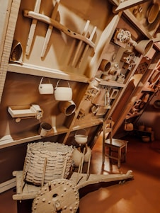 The image depicts a wall display of various rustic and vintage objects including metal pots, pans, a wooden cartwheel, a woven basket, and wooden tools, creating an antique and homely atmosphere.