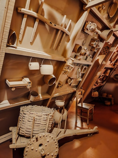 The image depicts a wall display of various rustic and vintage objects including metal pots, pans, a wooden cartwheel, a woven basket, and wooden tools, creating an antique and homely atmosphere.